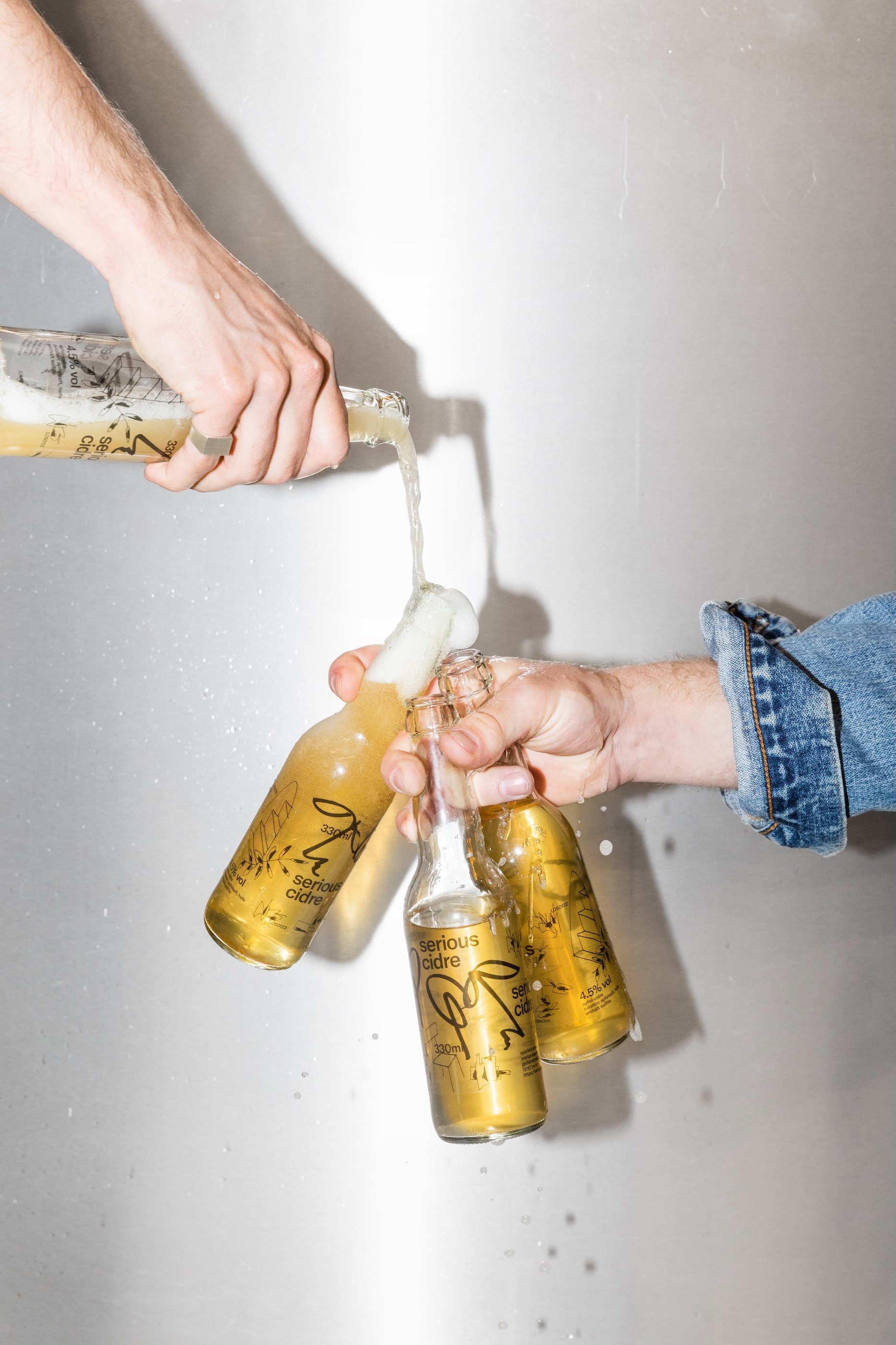 A photo of three overflowing bottles of Serious Cidre held by two hands, creating a mess.